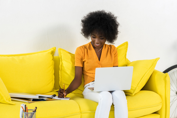 smiling african american female freelancer with laptop making notes while working on sofa at home - Photo, Image