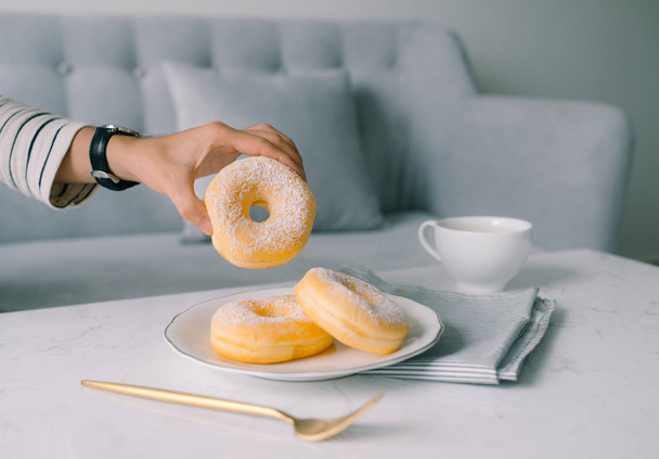 männliche Hand hält köstliche frische und süße Donuts mit Zuckerbelag mit Sofa auf dem Hintergrund. - Foto, Bild