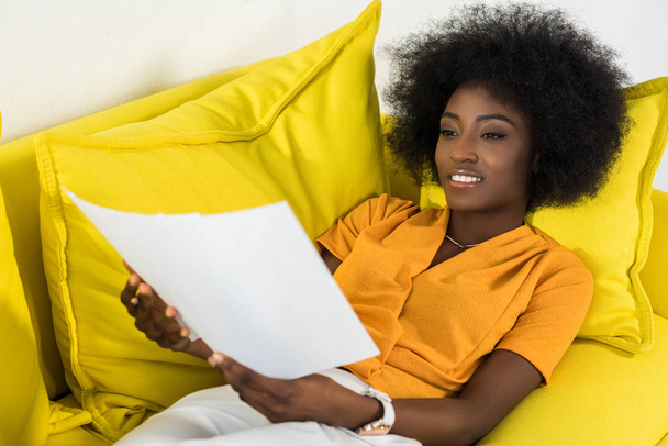 s úsměvem africká americká žena s doklady vzdálené pracuje na gauči doma - Fotografie, Obrázek