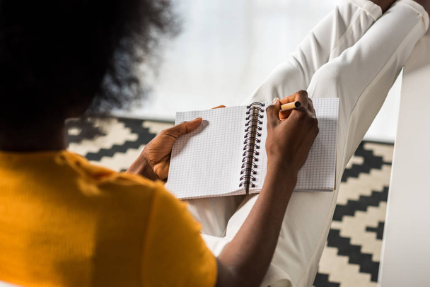 vista parcial del freelancer afroamericano haciendo notas en el cuaderno de la oficina en casa
 - Foto, Imagen