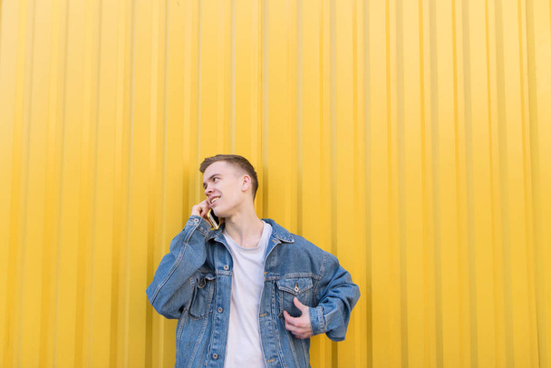 Un joven positivo con una chaqueta de mezclilla se encuentra en el fondo de una pared amarilla y habla por teléfono. Hablar por teléfono sobre un fondo amarillo
 - Foto, imagen