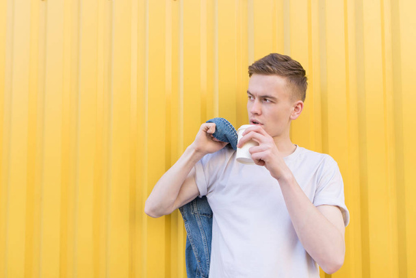 Giovane uomo in piedi su uno sfondo giallo, bere caffè e distogliere lo sguardo. Copyspace
. - Foto, immagini