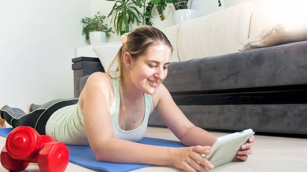 Mulher bonita deitada no chão na sala de estar e assistindo exercícios físicos em tablet PC
 - Foto, Imagem
