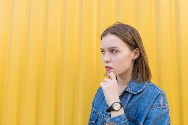 Une fille réfléchie se tient sur le fond d'un mur jaune et regarde sur le côté
 - Photo, image