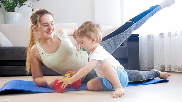 Joven mujer sonriente haciendo ejercicio físico mientras su hijo pequeño juega con juguetes a su lado
 - Foto, Imagen