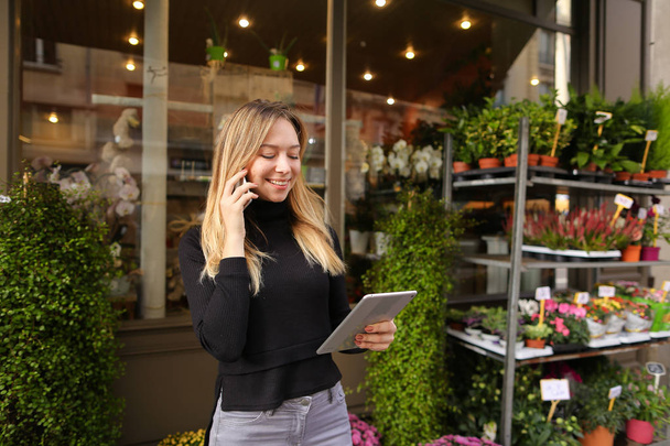 Женщина улыбается менеджер цветочного магазина стоя с планшетом и разговаривая по смартфону
. - Фото, изображение