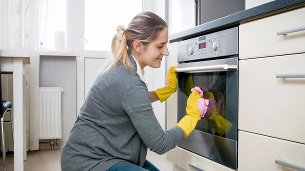Mosolygó fiatal nő Polírozó sütő üveg ajtó a konyhában - Fotó, kép