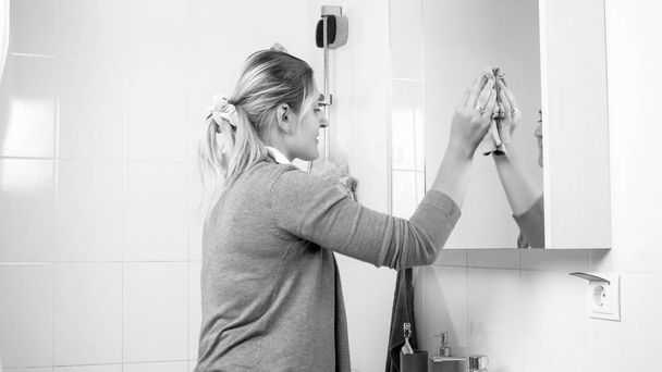 Fekete-fehér kép a fiatal háziasszony, tisztítás fürdőszoba tükör szennyeződéseket - Fotó, kép