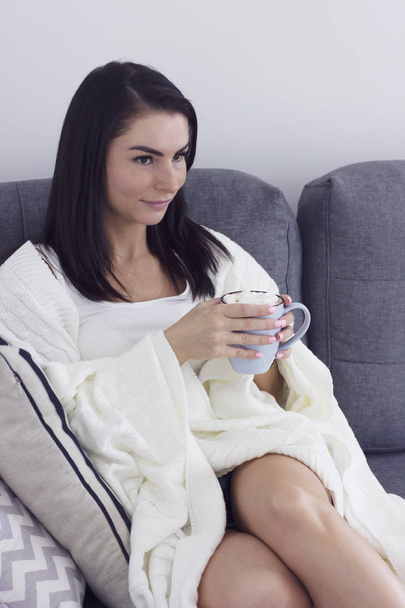 Mujer joven en manta con taza de cacao sentado en el sofá y viendo la televisión
 - Foto, Imagen