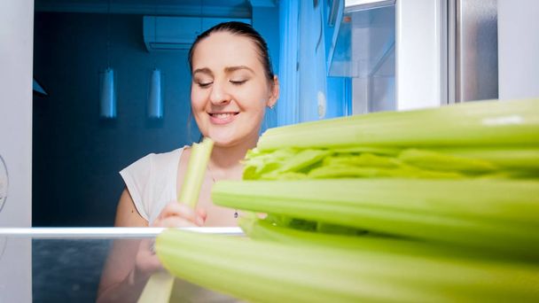 Closeup portrait of young woman eating celery she took from refrigerator. Concept of healthy eating - Photo, Image