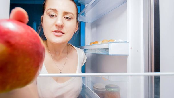 Nahaufnahme Porträt einer jungen Frau, die in den Kühlschrank schaut und roten Apfel nimmt - Foto, Bild
