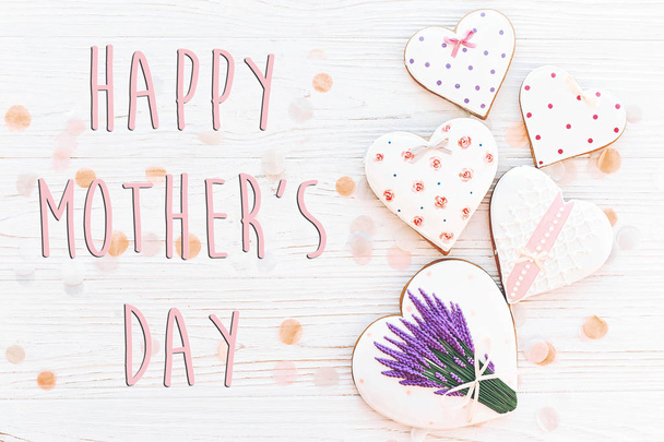 Gelukkig moeders dag tekst met roze cookies hartjes en paarse bloemen op witte rustieke houten achtergrond met confetti in zacht licht. - Foto, afbeelding