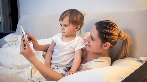 Portrait of smiling mother and toddler boy watching cartoons on tablet at bed - Photo, Image