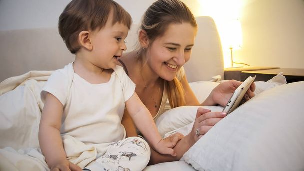 Closeup image of happy smiling mother with toddler son lying in bed and playing video game on digital tablet - Foto, imagen