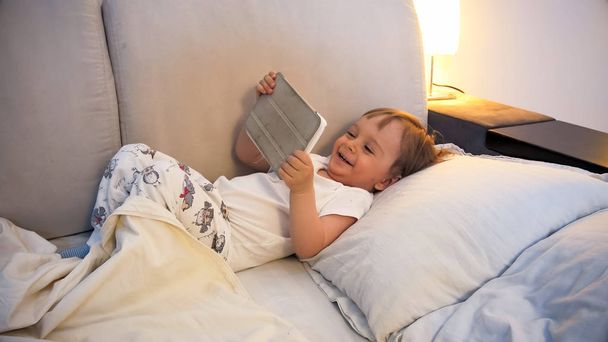 Portret szczęśliwy roześmiany toddler chłopiec leżąc w łóżku z cyfrowego tabletu - Zdjęcie, obraz