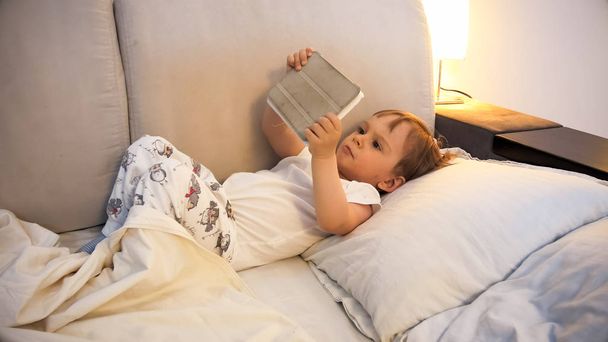 Portrait of cute toddler boy with digital tablet lying in bed - 写真・画像