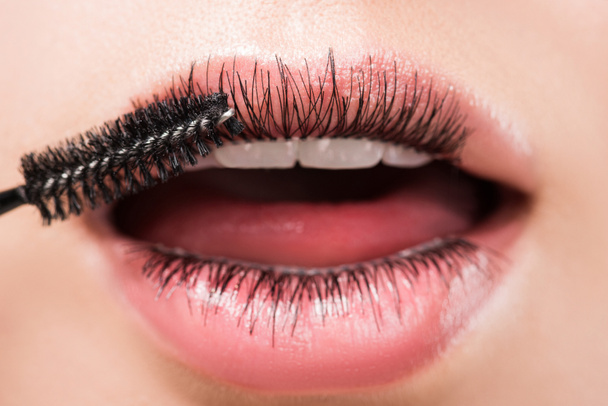 bijgesneden afbeelding van vrouw toepassing van zwarte mascara op wimpers in mond geïsoleerd op wit - Foto, afbeelding
