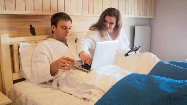 若い男の妻が付いているベッドで横になっていると、ノート パソコンを使用してのポートレート - 写真・画像