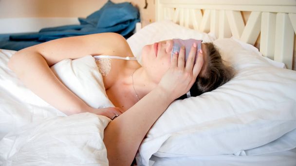 Closeup portret van een jonge vrouw met masker voor het slapen in bed liggen - Foto, afbeelding