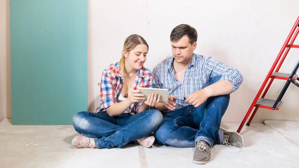 glückliches junges Paar sitzt im Bau befindlichen Haus auf dem Boden und nutzt digitales Tablet - Foto, Bild