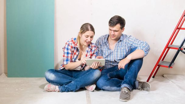 glückliches junges Paar sitzt im renovierten Haus auf dem Fußboden und surft auf einem digitalen Tablet im Internet - Foto, Bild