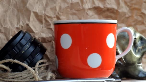 Steam rises over a cup of hot tea. Vintage red cup in white polka dots, twine, old lens from camera - Footage, Video