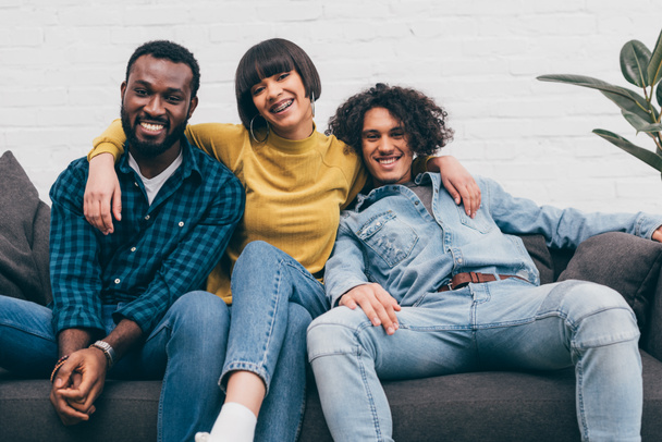 mixed race woman embracing two smiling male friends on couch - Photo, Image