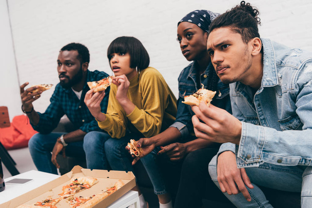 ryhmä nuoria monikansallisia ystäviä syö pizzaa ja katsomassa ottelua
 - Valokuva, kuva