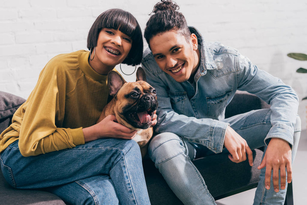 smiling young mixed race couple sitting on couch with french bulldog  - Zdjęcie, obraz