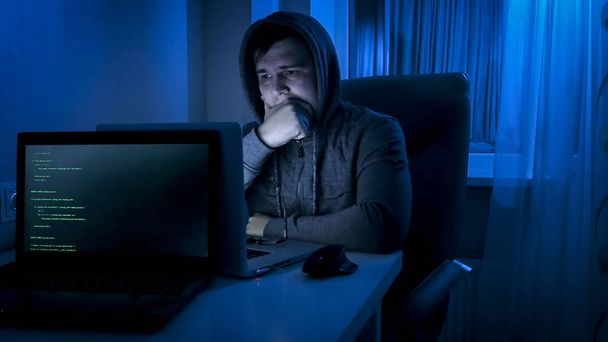 Νεαρός άνδρας στο hoodie ψάχνει laptop οθόνη τη νύχτα - Φωτογραφία, εικόνα