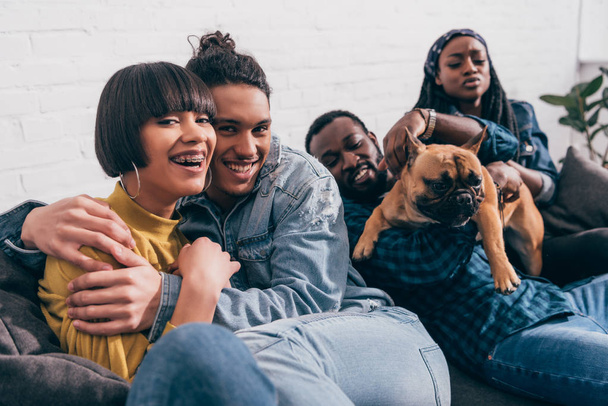 молодая улыбающаяся группа мультикультурных друзей, сидящих на диване с собакой
 - Фото, изображение
