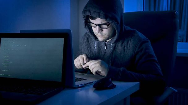Portrait of young man in hood holding credit card working on laptop - Photo, Image