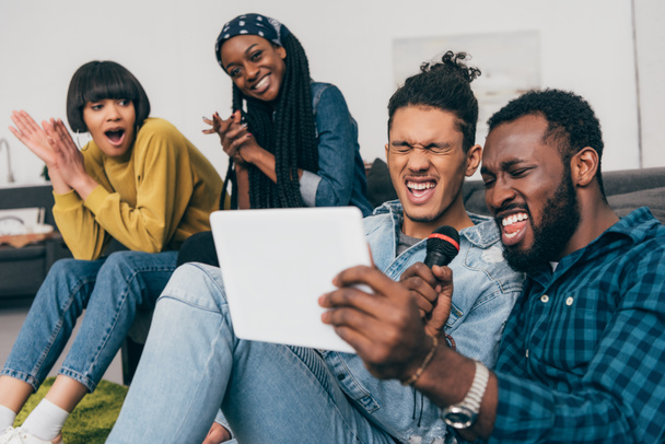 twee jonge multi-etnisch mannelijke vrienden met microfoon en digitale tablet zingen karaoke en vriendinnen zitten achter - Foto, afbeelding
