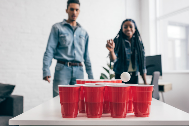 primer plano de la bola volando en taza de plástico en juego de pong cerveza y joven pareja multicultural de pie detrás
  - Foto, Imagen
