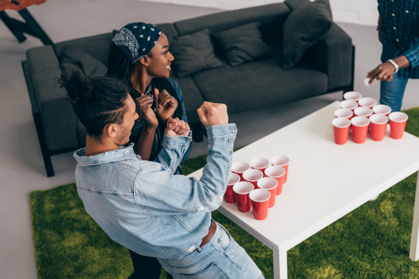 cropped shot of multicultural group of friends playing beer pong at table and young man doing winner gesture  - Photo, Image
