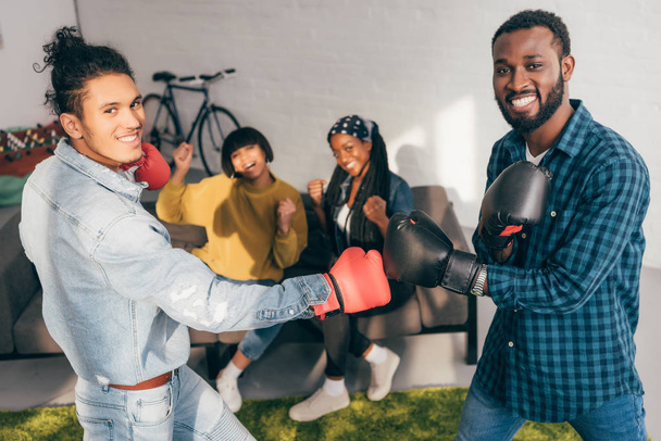 smiling multicultural male friends doing sparring in boxing gloves and two women cheering up them - Photo, Image