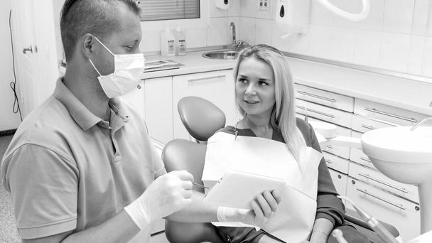 Zwart-wit beeld van tandarts praten met lachende jonge patiënt zit in de tandarts stoel - Foto, afbeelding