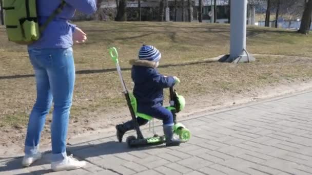 A young mother and a boy are walking outside. The kid skates sitting on the scooter - Footage, Video