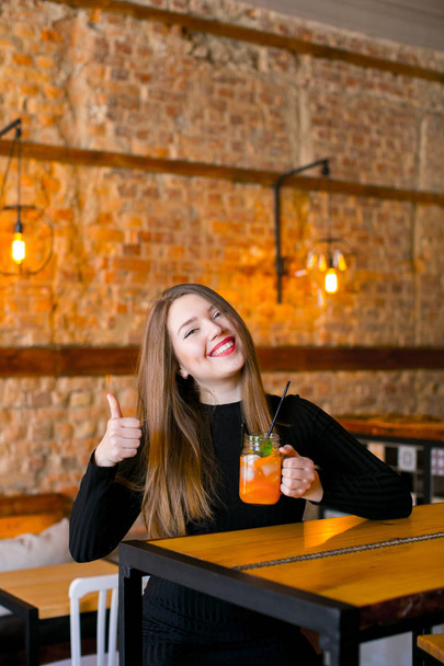 Tevreden meisje zit in de buurt van houten tafel met beker limonade in café en ontspannen. - Foto, afbeelding