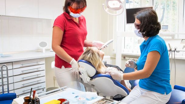 Женщина-ассистент помогает стоматологу при лечении зубов специальным ультрафиолетовым светом
 - Фото, изображение