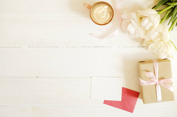 Femenine composition of woman's workspace w/ blank greeting card, envolope & presents for mother's day. Coffee cup with latte art & tulip flowe bouquet. Copy space, flat lay, background, top view. - Fotó, kép