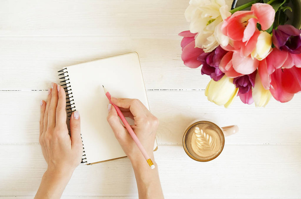 Overhead shot of woman hands drawing, writing with pencil in open notebook, drinking coffee on white wooden table. Beautiful tulip flowers bouquet of different colors. Background, copy space, close up - Valokuva, kuva