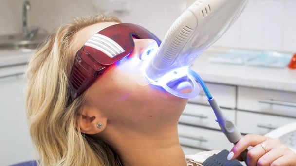 Portrait de jeune femme blonde dans une chaise de dentiste pendant le blanchiment des dents avec une lampe UV
 - Photo, image