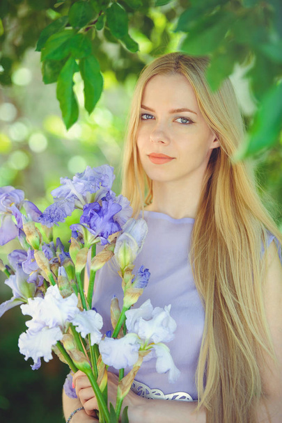 Attractive young girl with blonde hair and natural make-up smelling blue purple iris flowers on a background outdoors, tenderness and softness on a nature background - Foto, afbeelding