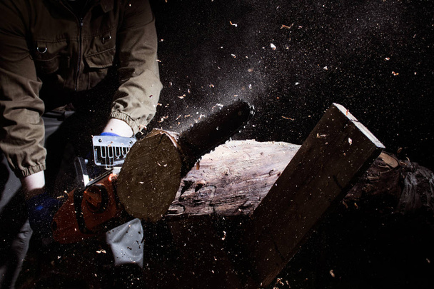 Foto de un trabajador vestido con guantes azules cortando troncos de madera sobre fondo negro
. - Foto, Imagen