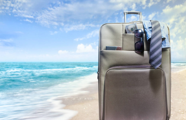 Φωτογραφία: μια βαλίτσα ταξιδίου συσκευάζονται με πουκάμισο, γραβάτα, χρήματα και γυαλιά ηλίου που στέκεται πάνω σε ένα φόντο παραλία sunny. - Φωτογραφία, εικόνα