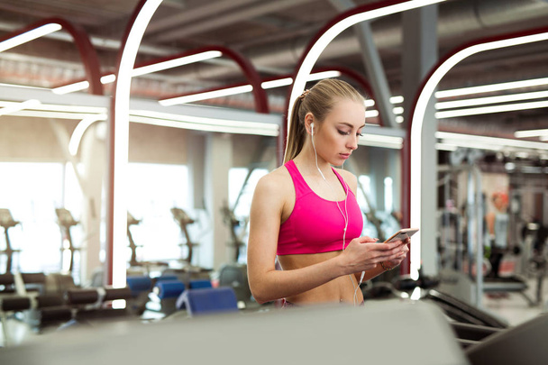 a beautiful young girl in a pink top is engaged in walking on a treadmill with a phone in her hands - Photo, Image