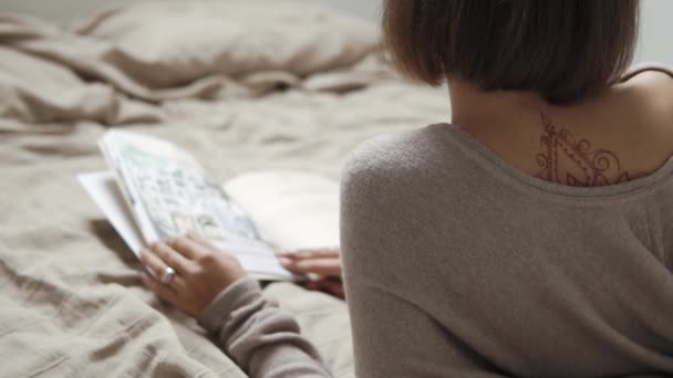 Achteraanzicht van slanke eenzame vrouw met tatoeage liggend in bed met boek - Video