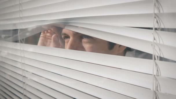 Молодой человек смотрит в окно через жалюзи на улицу
 - Кадры, видео