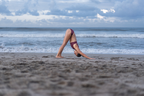 side view of attractive young woman practicing yoga in Downward-Facing Dog pose (Adho Mukha Svanasana) on seashore - Fotoğraf, Görsel